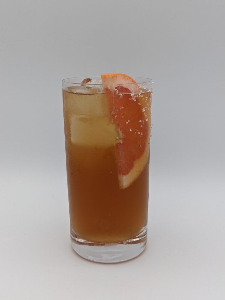brownish orange liquid in a tall glass with large ice cubes, grapefruit slice garnish and salted rim on the right half