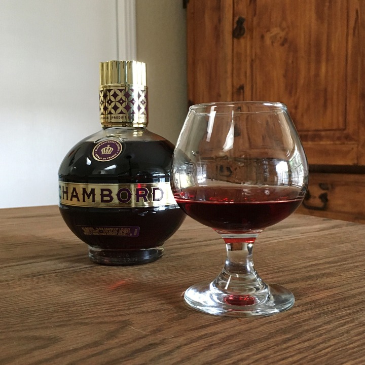 Bottle of Chambord Black Raspberry Liqueur next to a rounded glass filled with dark purple liquid, sitting on a wooden table