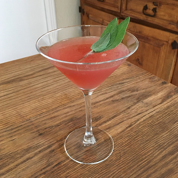 bright pink-red cocktail in a martini glass with sage leaf garnish, sitting on a wooden table
