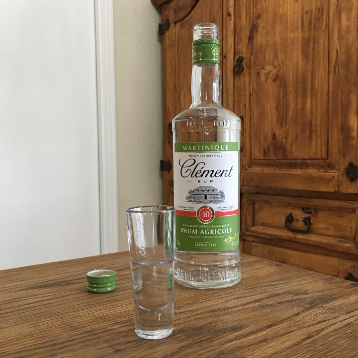 Open bottle of Clément Rhum Agricole Blanc next to a tall shot glass with clear liquid, both sitting on a wooden table