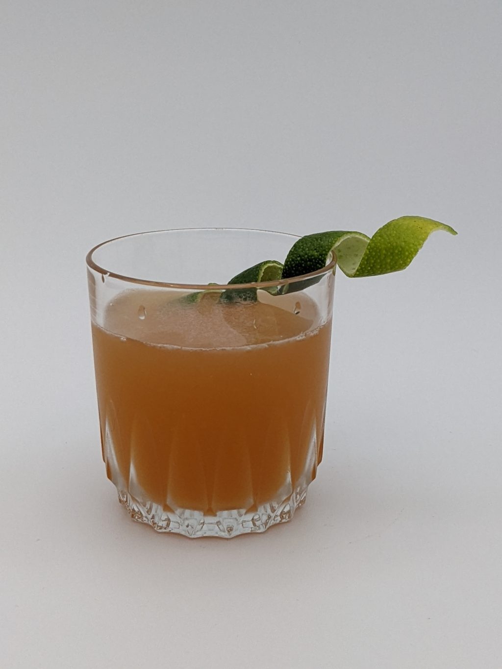 orange and brown liquid in a clear glass with a curled lime peel as a garnish