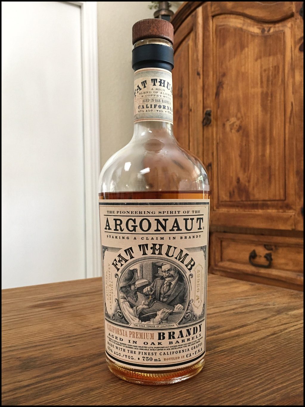 Bottle of Argonaut Fat Thumb Brandy sitting on a wooden table, in front of a mixed white and wooden background
