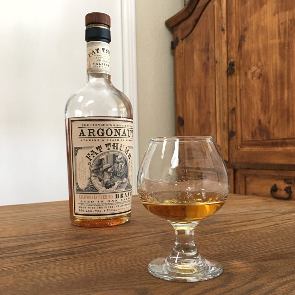 Bottle of Argonaut Fat Thumb Brandy next to a small rounded glass with golden liquid, both sitting on a wooden table