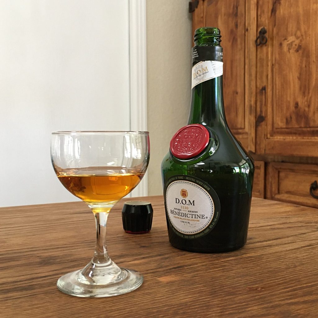 Open bottle of Benedictine D.O.M Liqueur, sitting behind a small stemmed cocktail glass with golden liquid