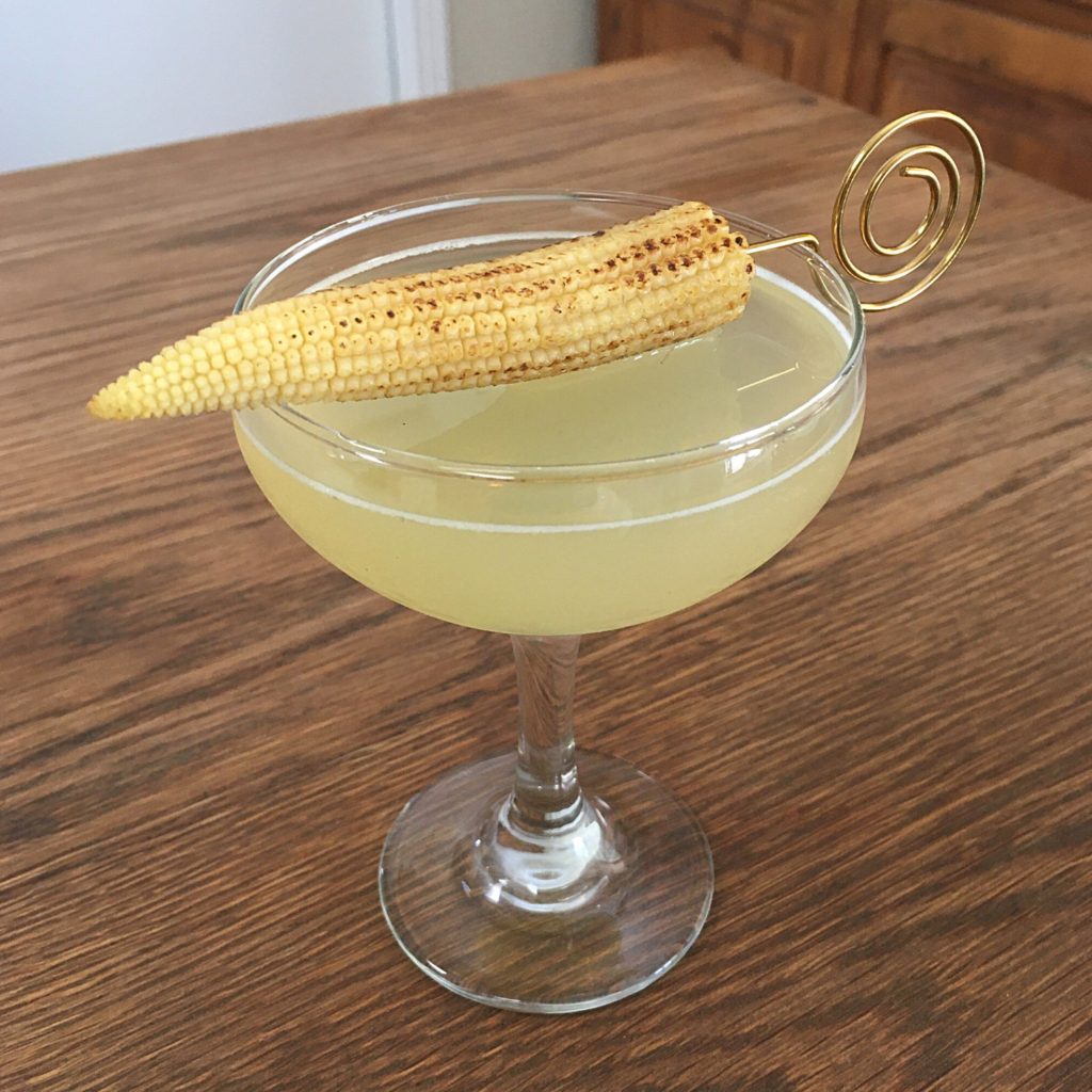 Light yellow cocktail in a coupe glass with a charred baby corn garnish arranged with a golden cocktail pick