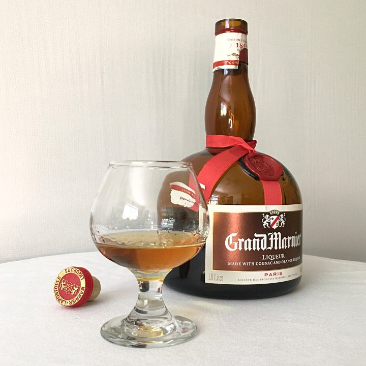 Open bottle of Grand Marnier sitting next to a small glass of light amber liquid and a cork cap, all sitting on a white tablecloth