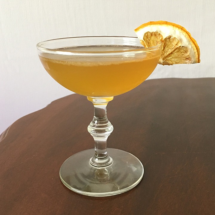 Orange-yellow cocktail in a coupe glass with dried lemon triangle garnish, sitting on a wooden table