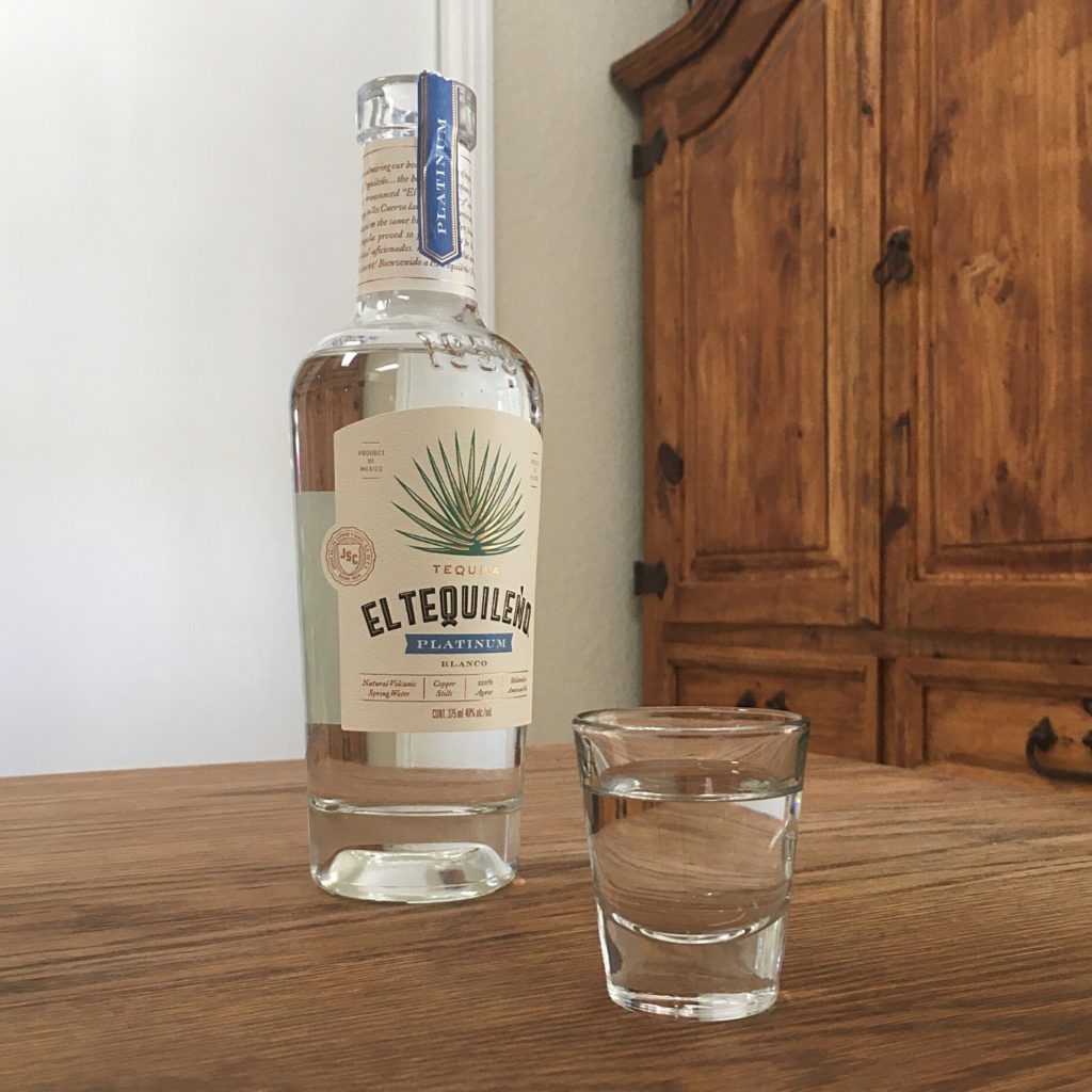 Open Bottle of Tequileño Platinum Blanco Tequila next to a shot glass full of clear liquid, both sitting on a wooden table