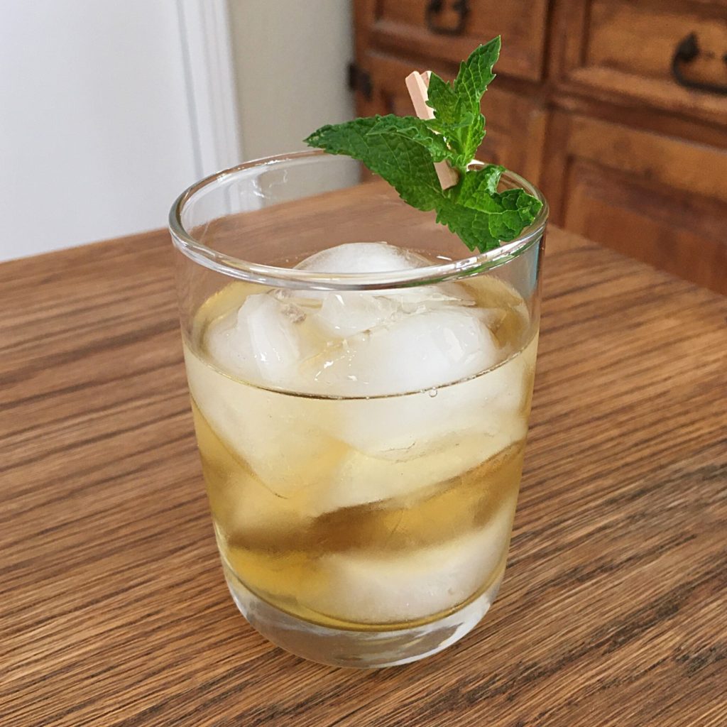 Light golden cocktail with ice and a mint sprig garnish, sitting on a wooden table