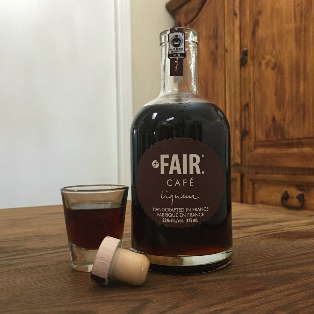 Open bottle of Fair Cafe Liqueur sitting next to its cork and a shot glass of dark brown liquid, all sitting on a wooden table
