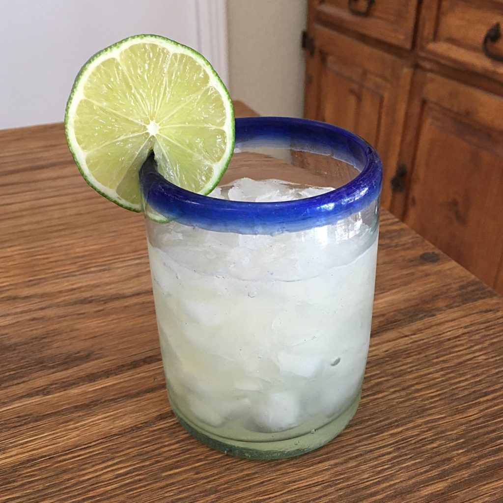 Margarita cocktail in small hand-blown glass with a blue rim and a lime wheel garnish, sitting on a wooden table