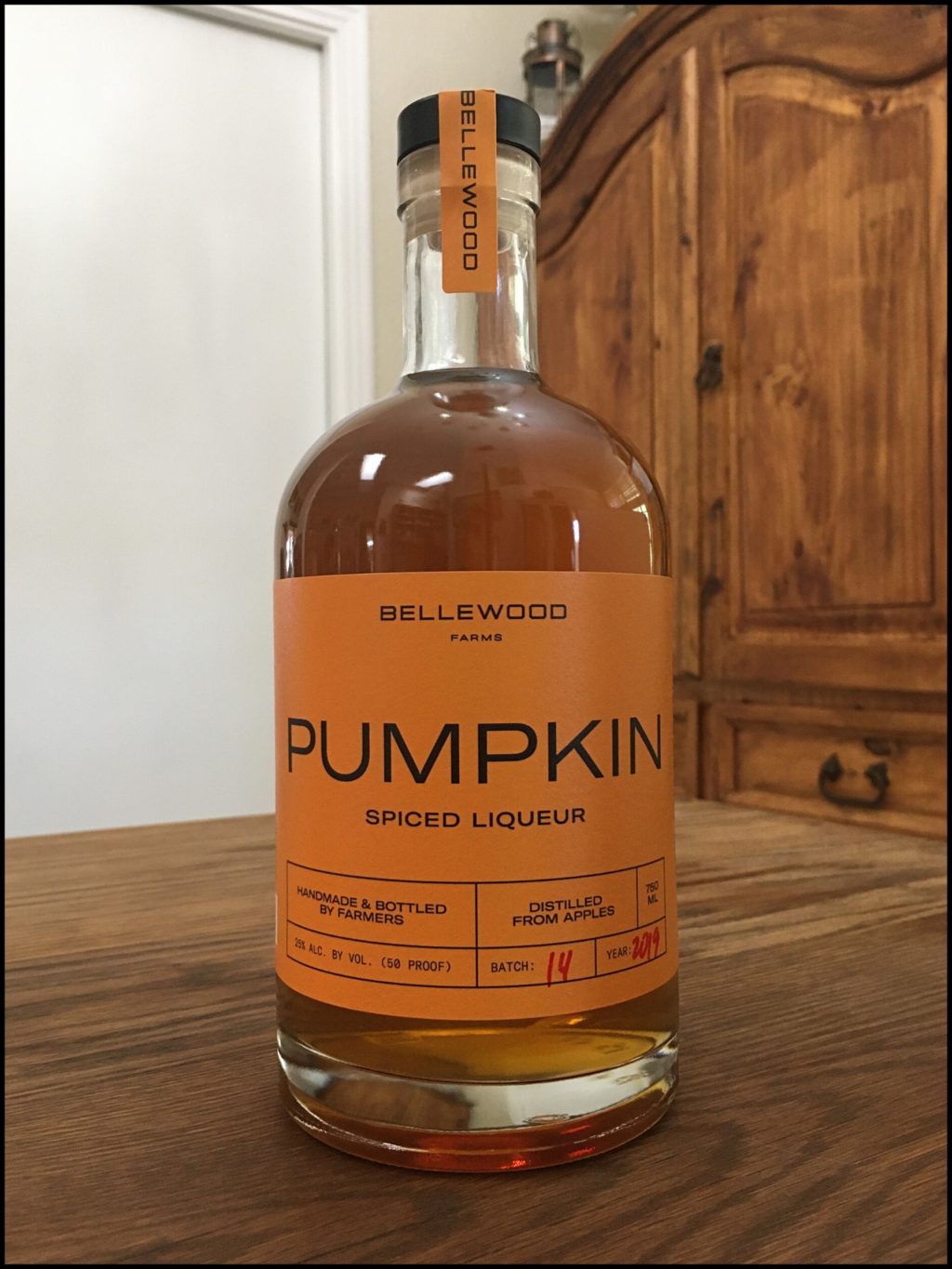 Bottle of Bellewood Farms Pumpkin Spice Liqueur sitting on a wooden table, in front of a mixed white and wooden background