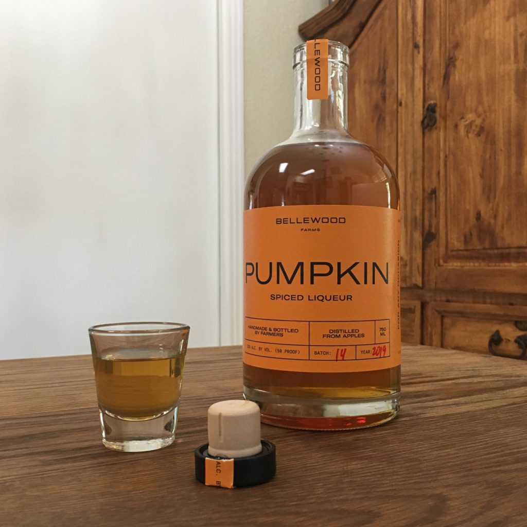 Open bottle of Bellewood Farms Pumpkin Spice Liqueur sitting next to its cork cap and a shot glass filled with brown-orange liquid, all sitting on a wooden table