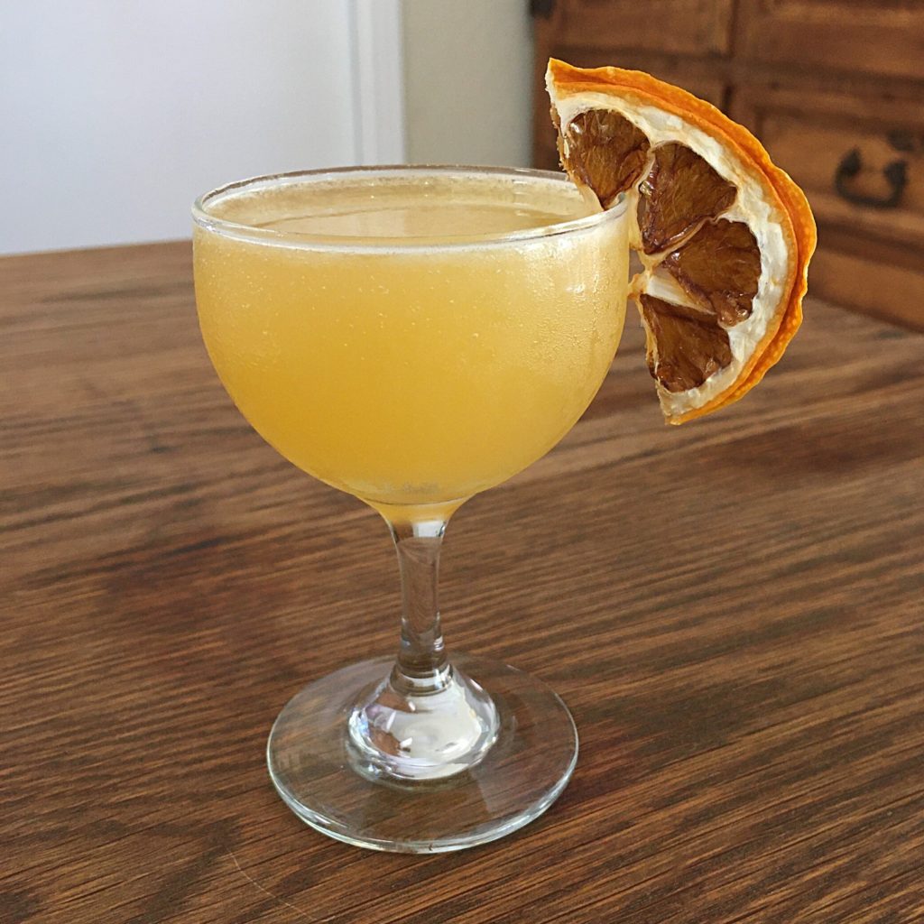 Small round stemmed glass filled with orange-yellow liquid and a dried lemon slice garnish