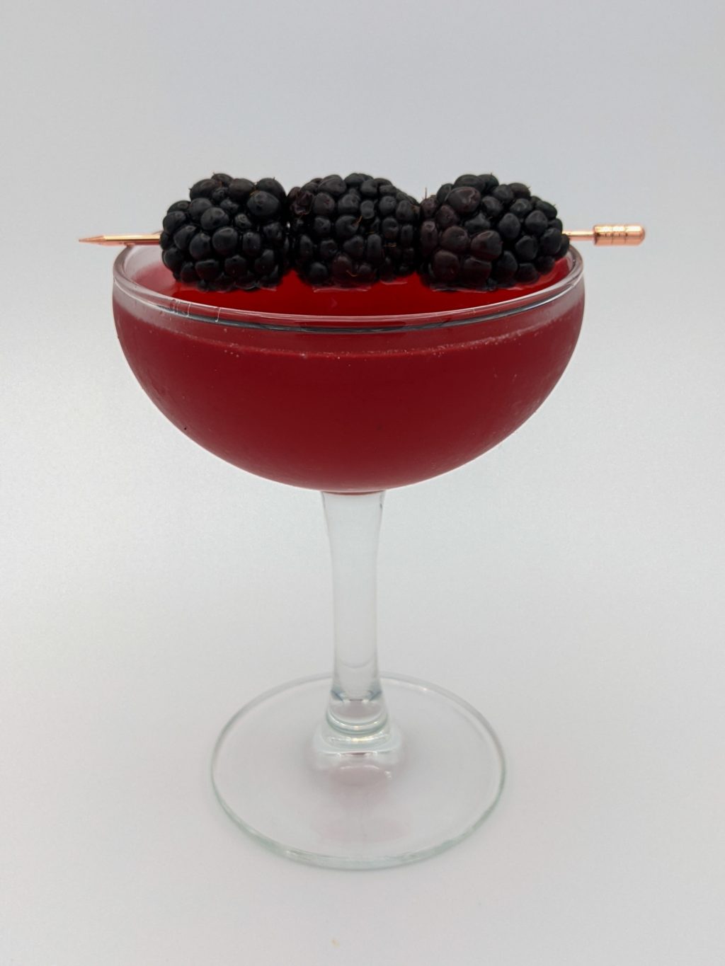 reddish purple liquid in a coupe glass with three black berries skewered with a gold cocktail pick