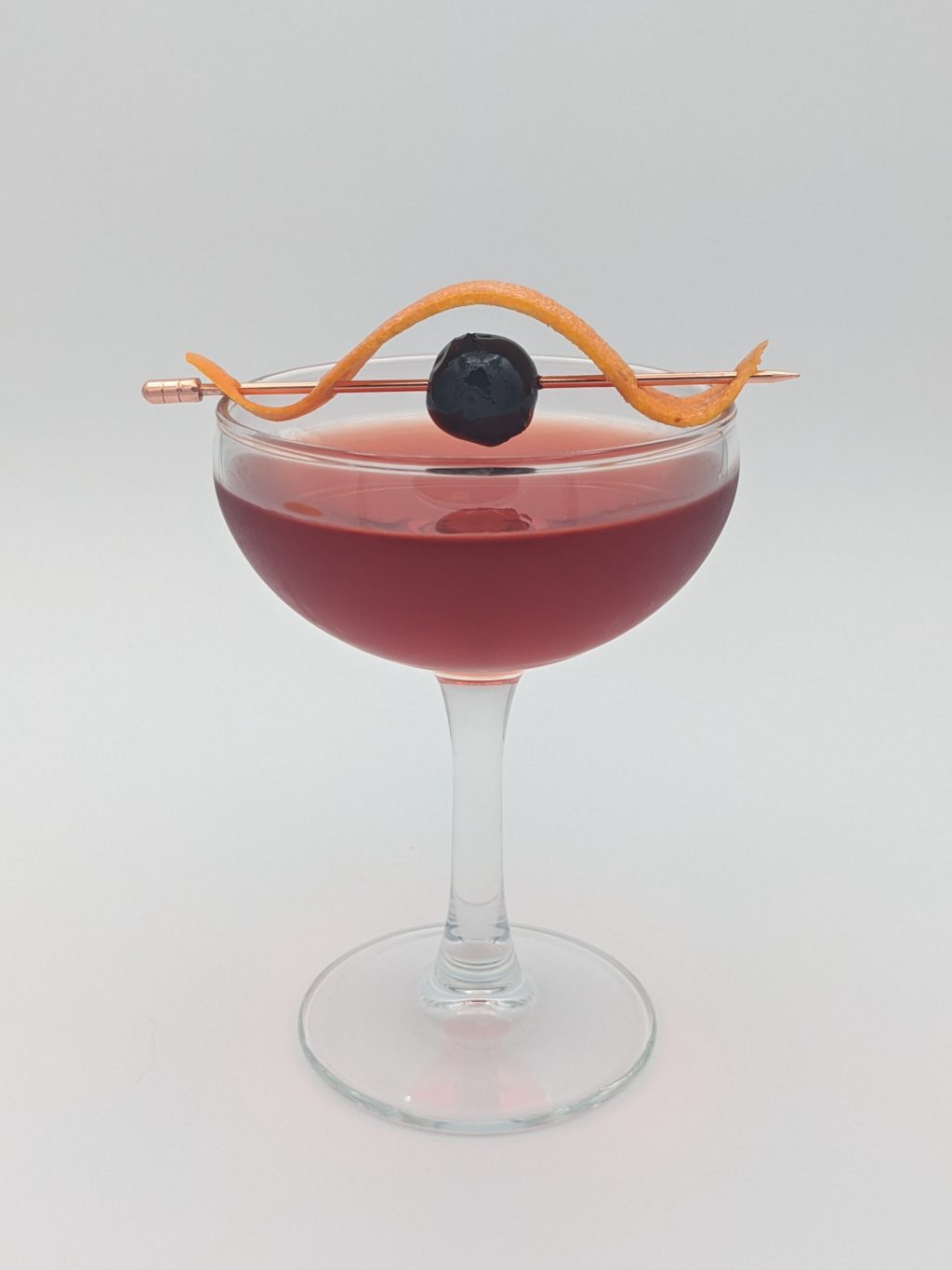 dark red liquid in a coupe glass with a orange peel and cherry garnish
