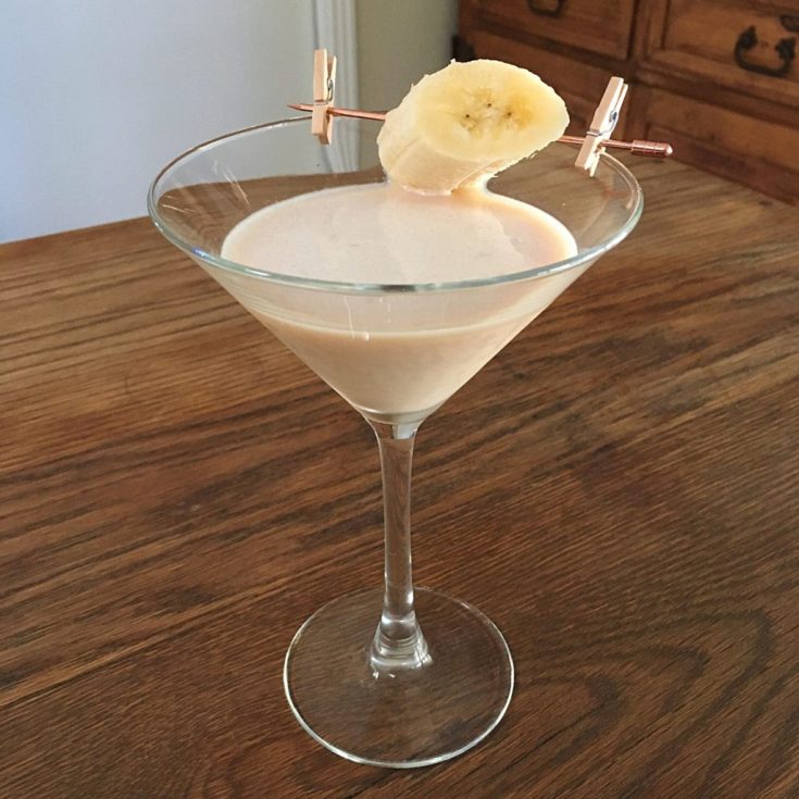 Creamy beige liquid in a martini glass with banana slice garnish that is attached with a rose goldtone cocktail pick and two clothespins