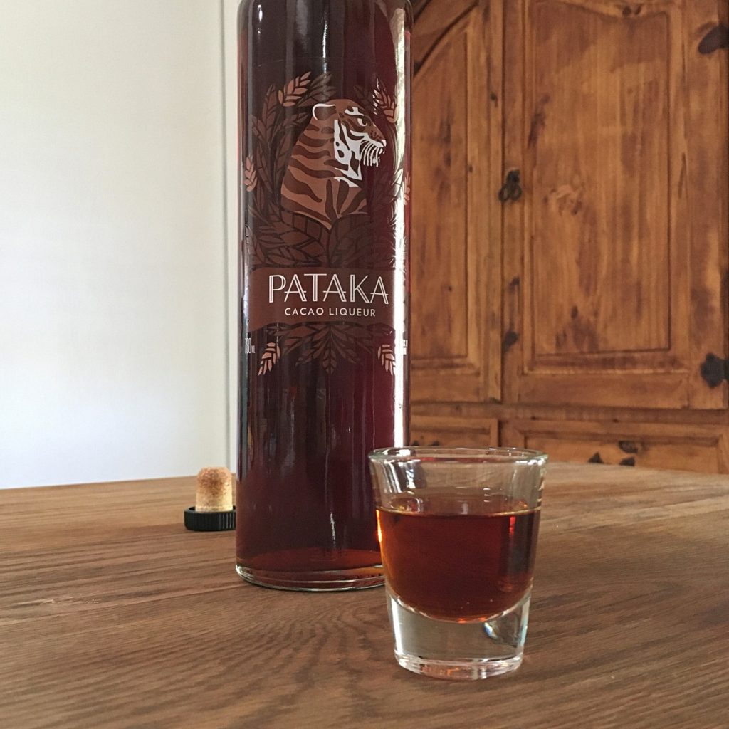 Mid close up of a shot glass filled with brown liquid, with a tall bottle partially showing behind it with the "Pataka Cacao Liqueur" label visible