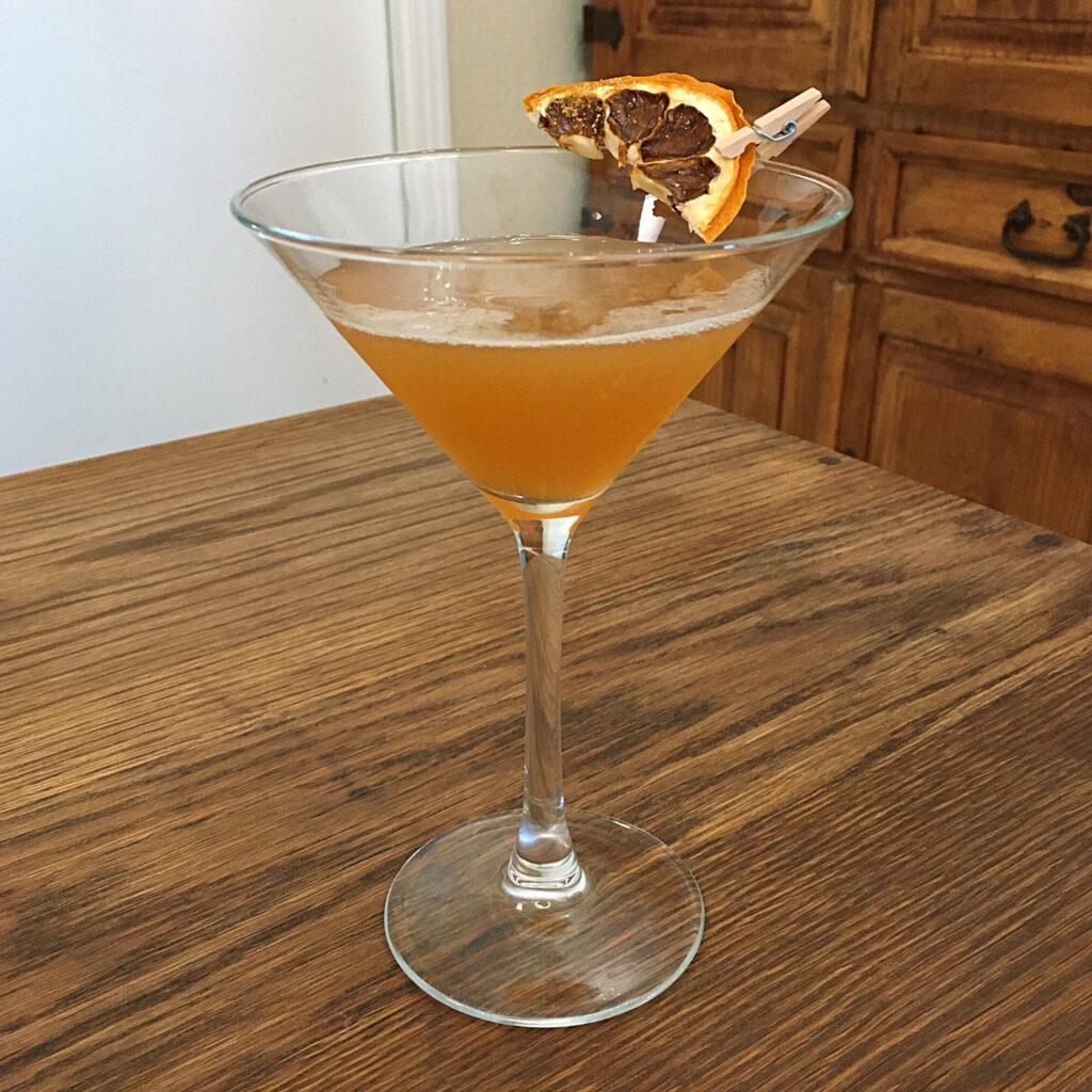Martini glass full of golden liquid sitting on a wooden table. The rim of the glass is garnished with a dried lemon slice sprinkled with edible gold glitter and pinned with an extra small clothespin.