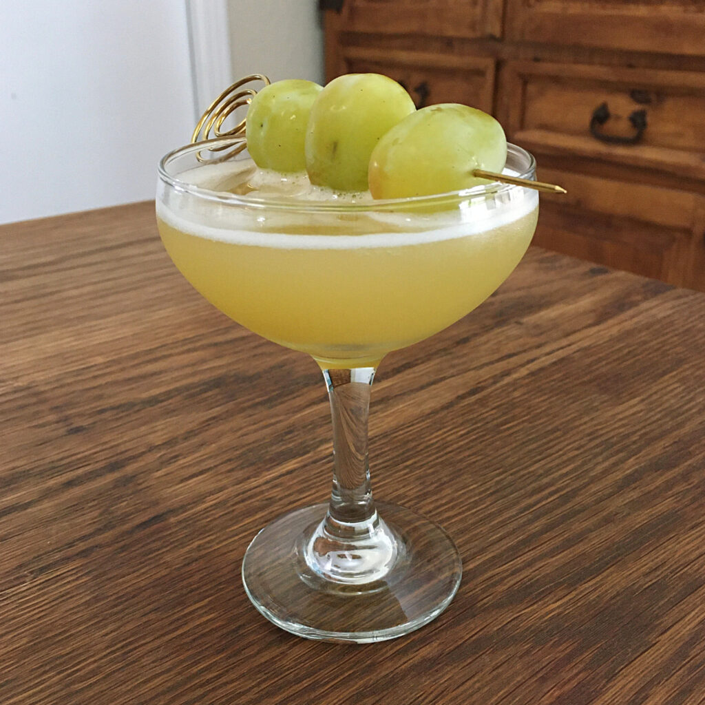 greenish yellow cocktail in a coupe glass garnished with three large green grapes skewered on a goldtone cocktail pick
