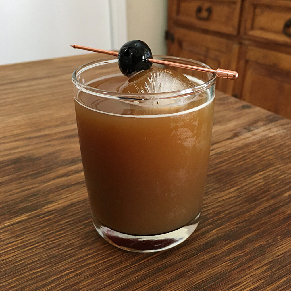 Golden brown cocktail with one large round ice cube and a maraschino cherry garnish on a rose gold cocktail pick, sitting on a wooden table