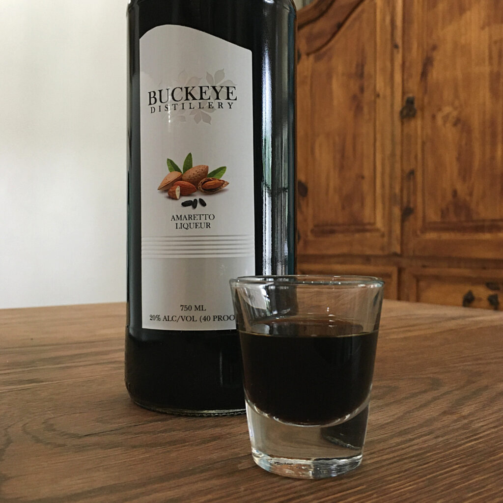 Bottle of Buckeye Amaretto Liqueur with just the label showing, sitting next to a shot glass filled with dark purple brown liquid, both sitting on a wooden table