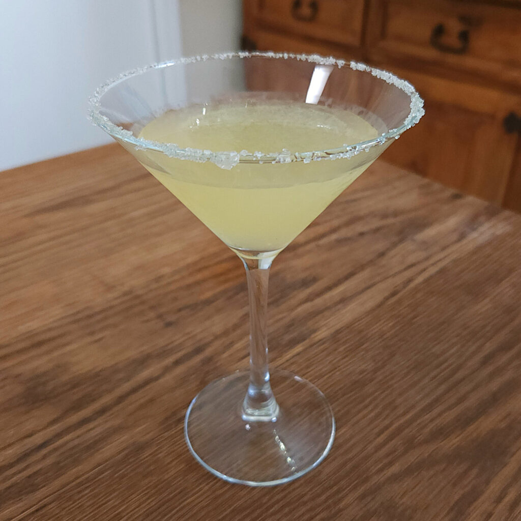 light yellow cocktail in a martini glass and sugar rim garnish, sitting on a wooden table