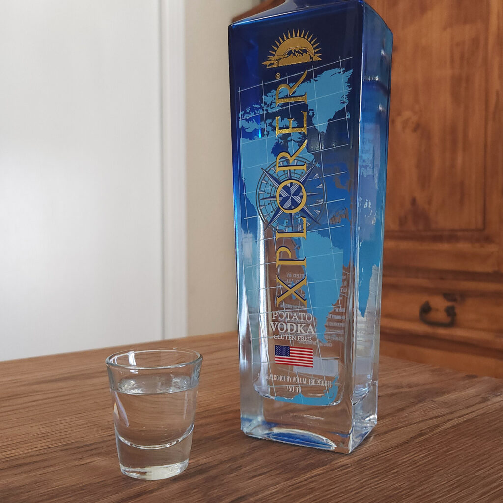 Bottle of Xplorer Potato Vodka next to a shot glass filled with clear liquid, both sitting on a wooden table