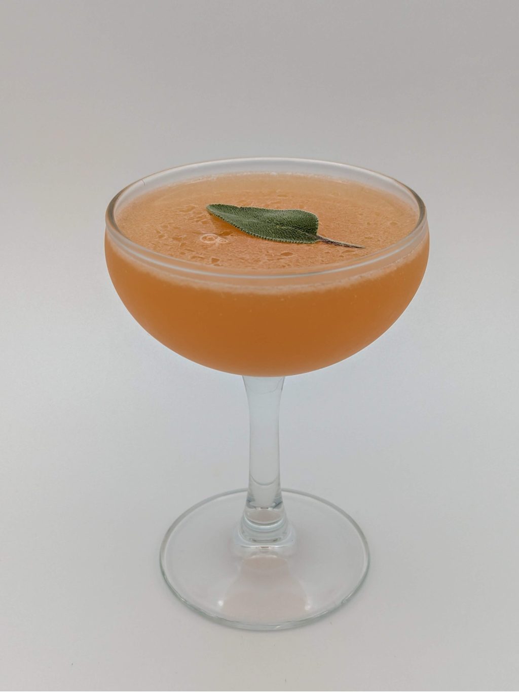 orange liquid in a coupe glass with a sage leaf floating on top