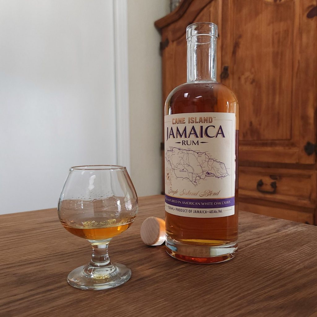 Open bottle of Cane Island Jamaica Rum slightly in front of a cork cap, and slightly behind a snifter glass partially full of golden liquid, all sitting on a wooden table