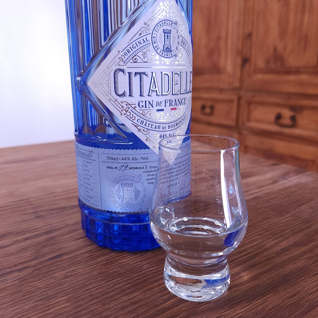 Bottle of Citadelle Gin with half the bottle showing, next to a small snifter glass with clear liquid, both sitting on a wooden table