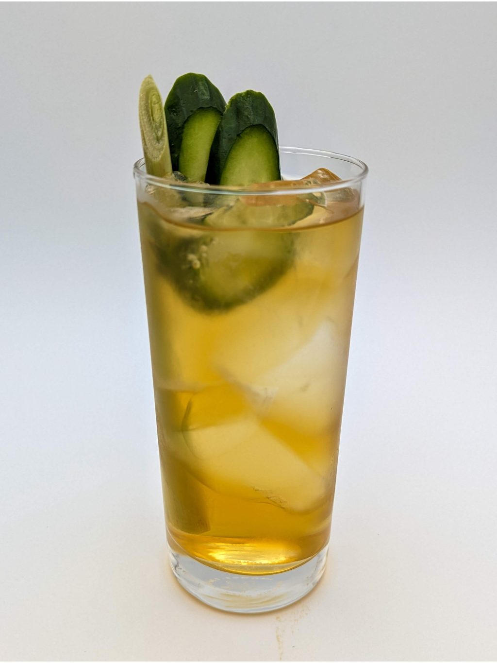 Golden Liquid in a tall glass filled with ice and garnished with a lemongrass stalk and two slices of cucumber