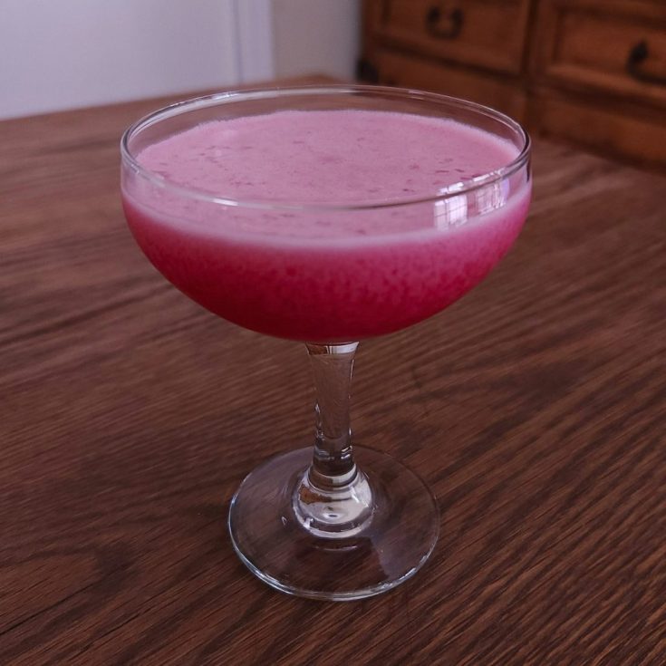 Coupe glass with an opaque pink-red cocktail with slight white foam, sitting on a wooden table