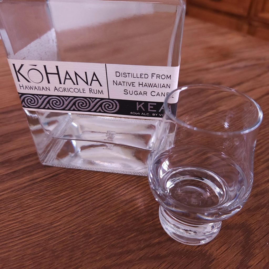 Bottle of Ko Hana Hawaiian Agricole Rum with the bottom half of the bottle showing, sitting next to a small snifter glass with clear liquid, both sitting on a wooden table