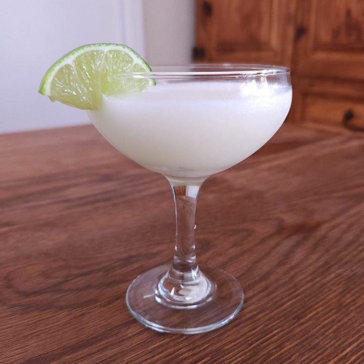 Stemmed cocktail glass with opaque white liquid and a lime slice garnish, sitting on a wooden table