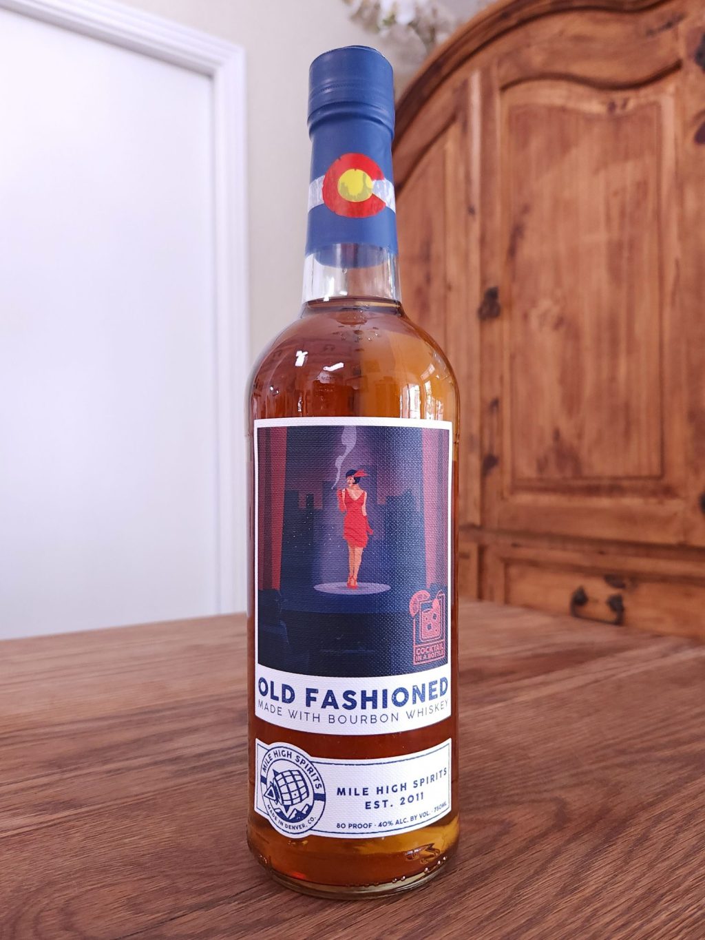 Bottle of Fireside Old Fashioned sitting on a wooden table, in front of a mixed white and wooden background
