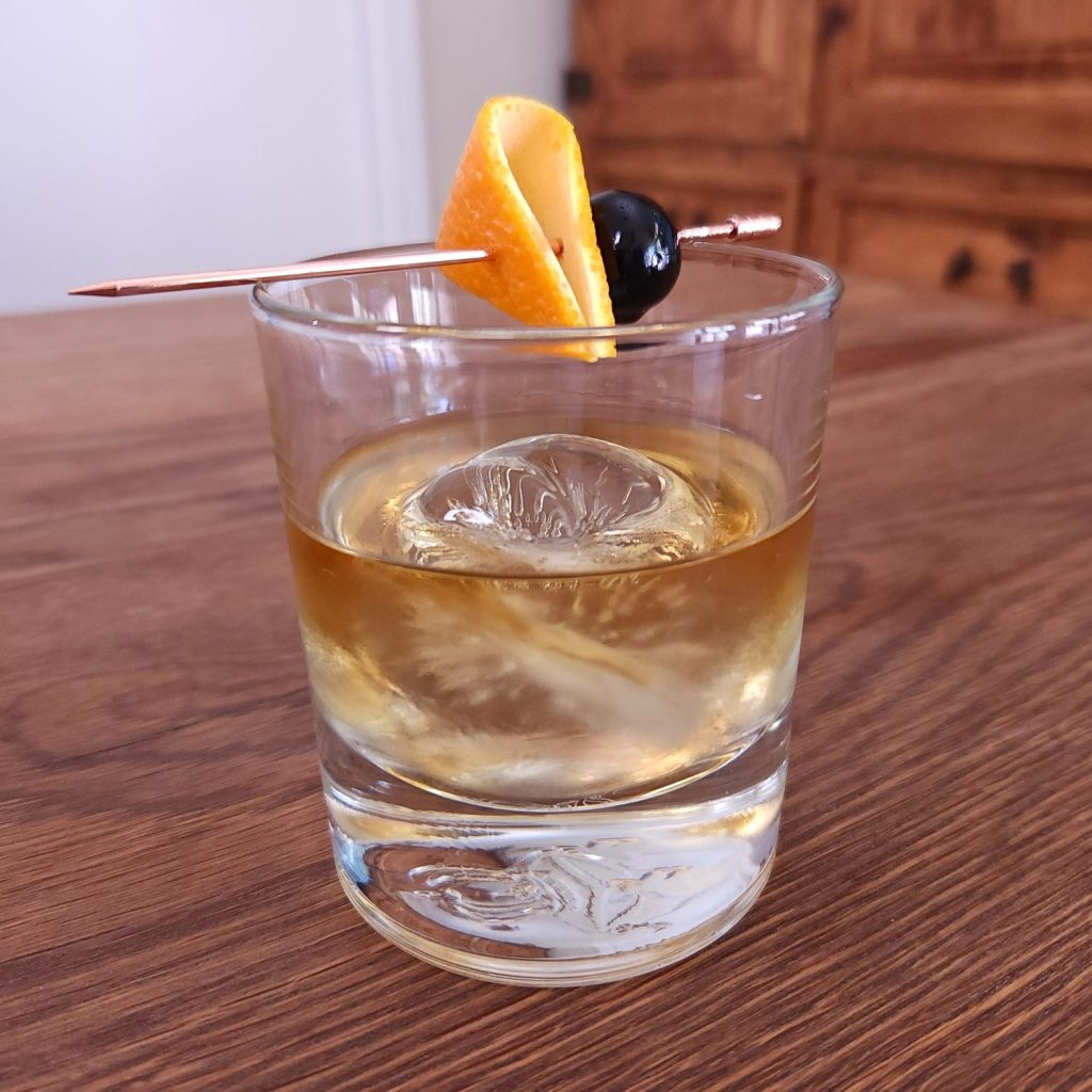 Old Fashioned glass with light amber liquid, one ice sphere, and a rose gold cocktail pick with orange peel and cherry garnish. The glass is sitting on a wooden table
