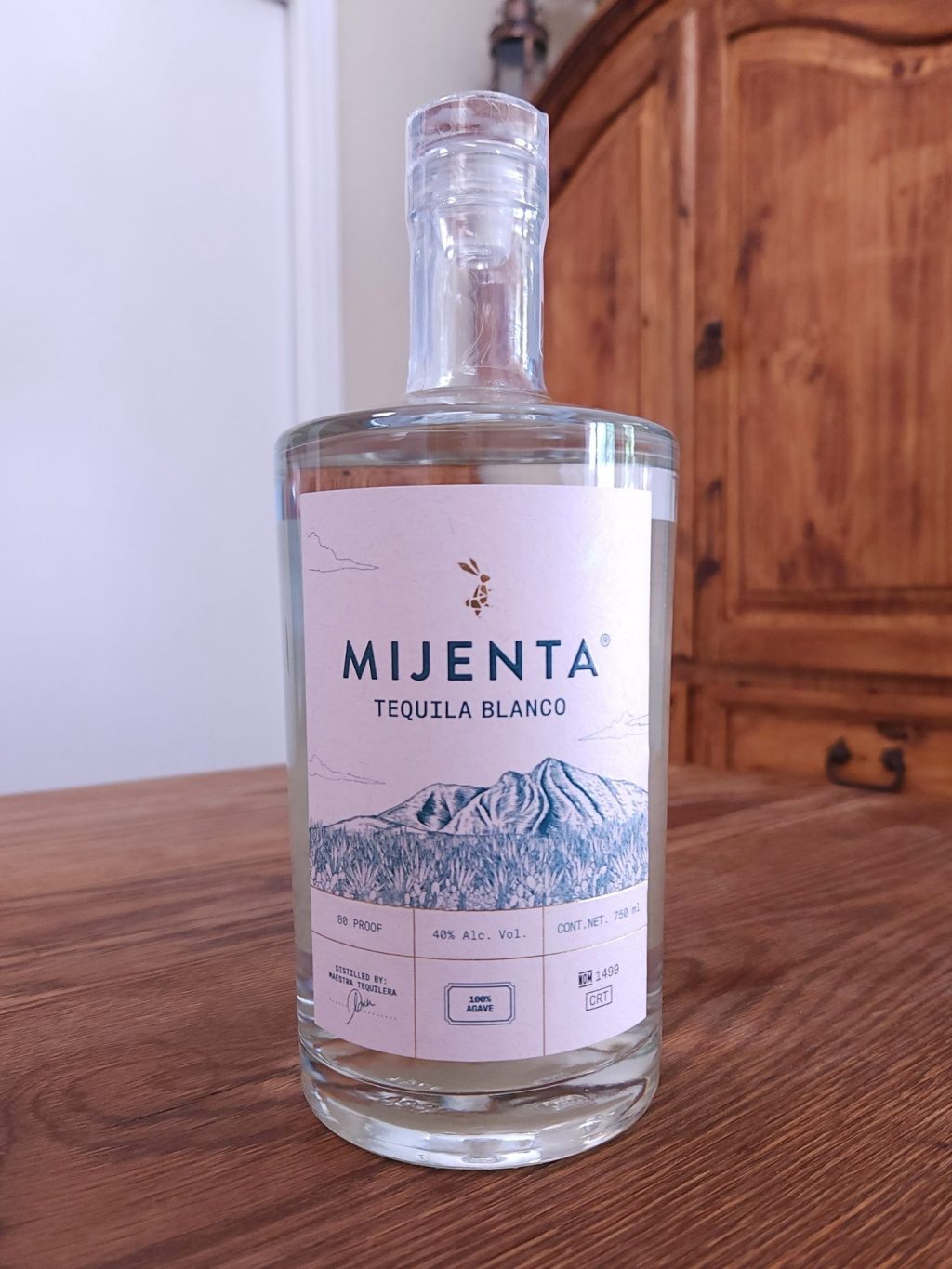 Bottle of Mijenta Blanco Tequila sitting on a wooden table, in front of a mixed white and wooden background.