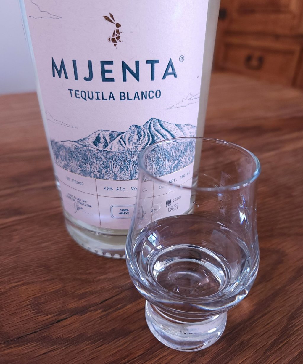 Small snifter glass with clear liquid, next to a bottle of Mijenta Blanco Tequila with only the label showing, both sitting on a wooden table
