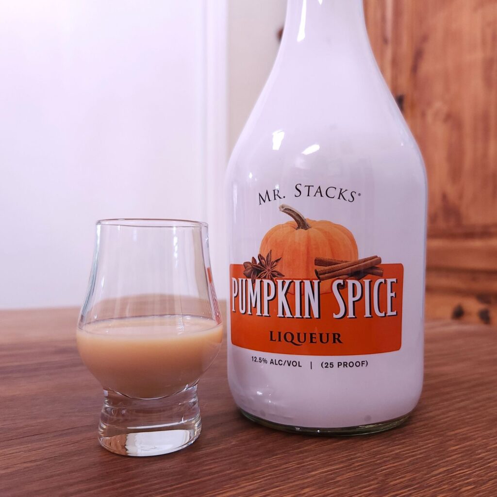 Bottle of Mr Stacks Pumpkin Spice Liqueur with only the label showing, next to a small snifter glass with opaque creamy beige liquid, both sitting on a wooden table