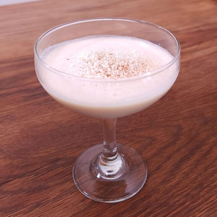 Stemmed coupe glass sitting on a wooden table and filled with creamy liquid, sprinkled with white chocolate shavings and golden brown powdered spice on the top
