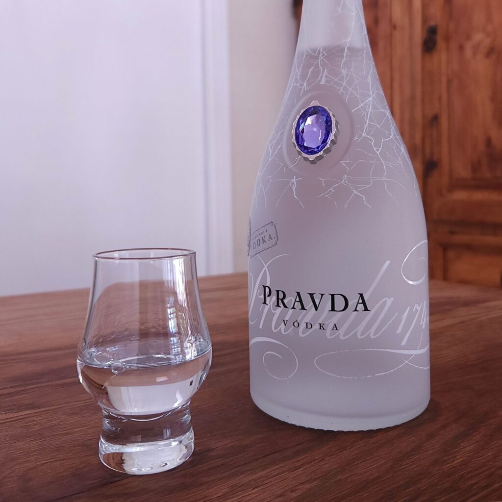 Small snifter glass with clear liquid sitting next to a bottle of Pravda Vodka with only the label showing, both sitting on a wooden table