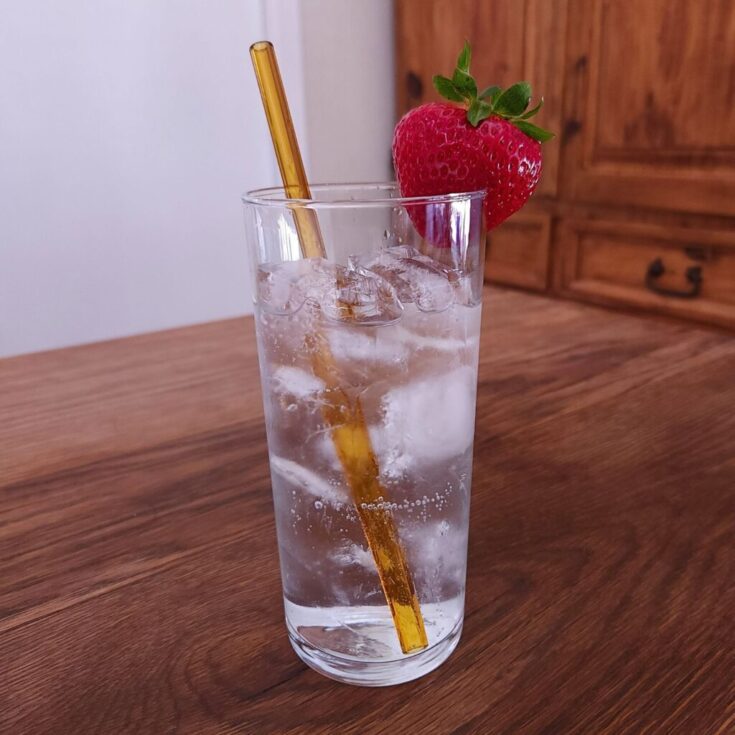Tall glass filled with ice and clear carbonated liquid, with an amber yellow glass straw and whole strawberry garnish