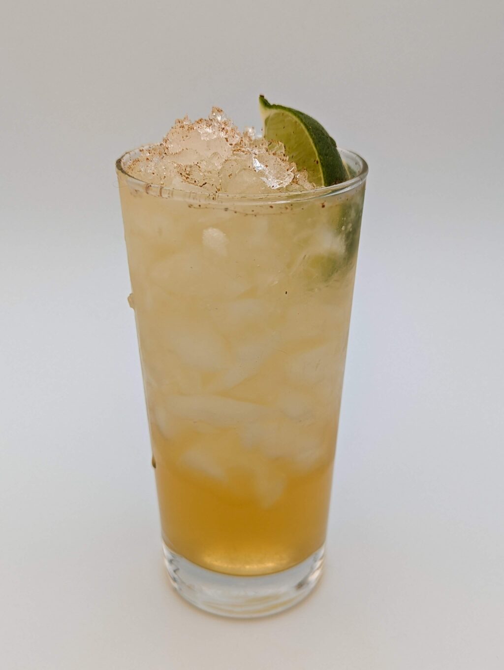 gold liquid in a tall glass filled with crushed ice with a lime wedge garnish