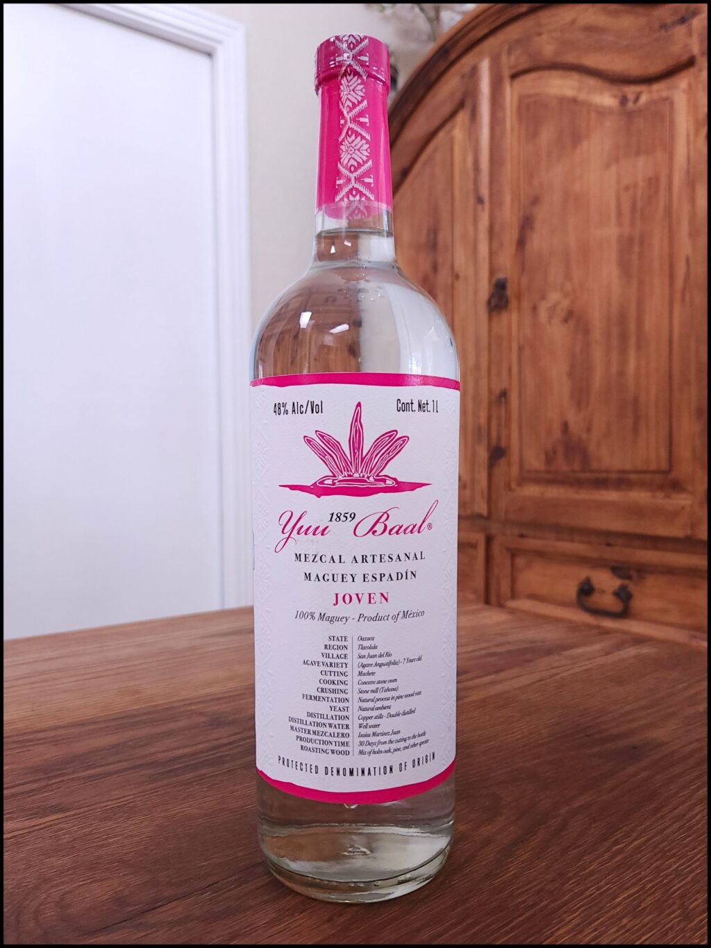 Bottle of Yuu Baal Mezcal Joven sitting on a wooden table, in front of a mixed white and wooden background