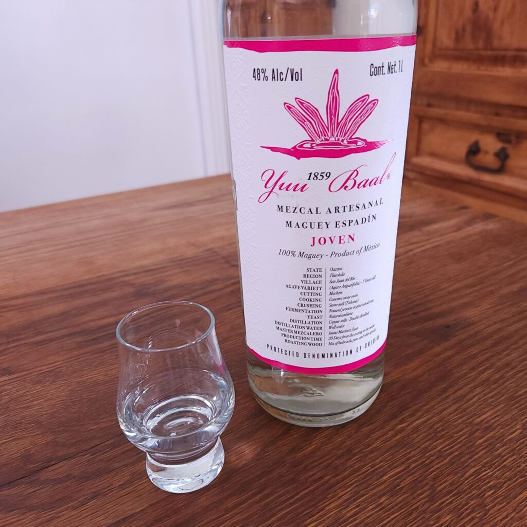 Small snifter glass with clear liquid next to a bottle of Yuu Baal Mezcal Joven with only the label showing, both sitting on a wooden table