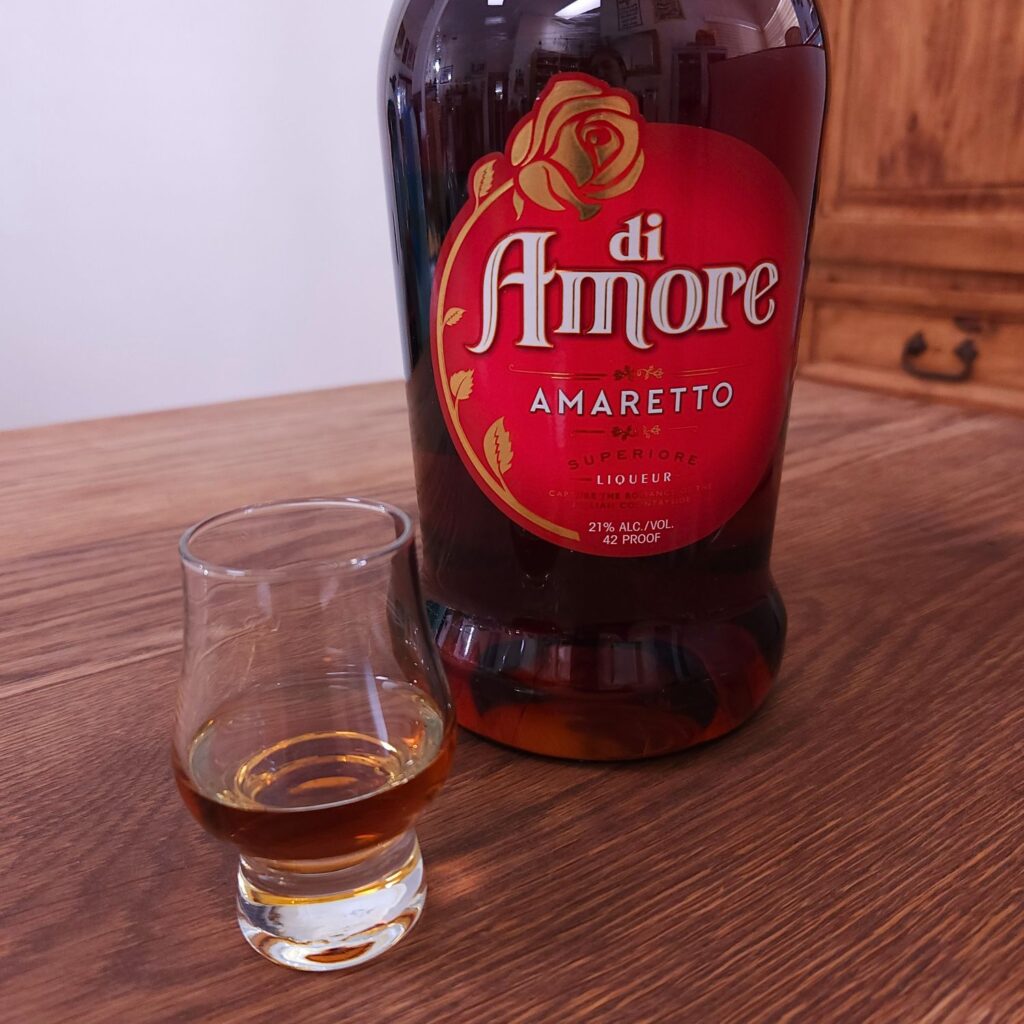 bottle of Di Amore Amaretto with only the bottom half showing, next to a small snifter glass with dark amber liquid, both sitting on a wooden table