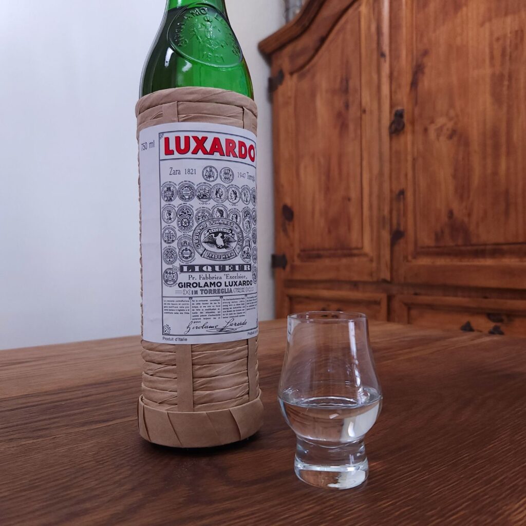 Bottle of Luxardo Maraschino Liqueur with only the label showing, next to a small snifter glass with clear liquid, both sitting on a wooden table