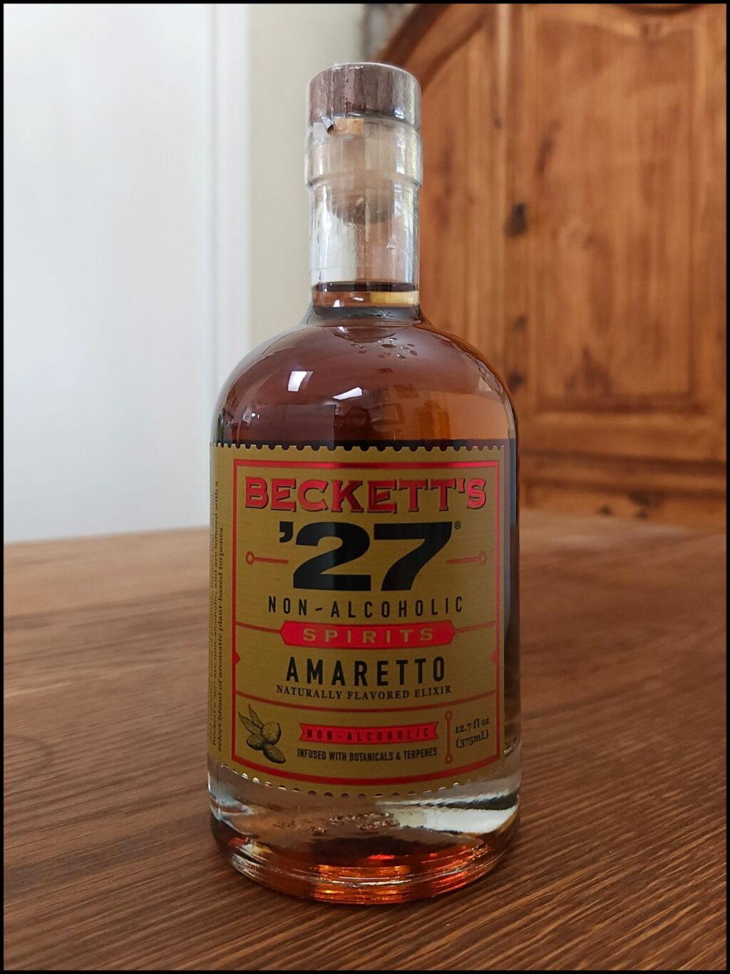 Bottle of Beckett's '27 Non-Alcoholic Amaretto sitting on a wooden table, in front of a mixed white and wooden background