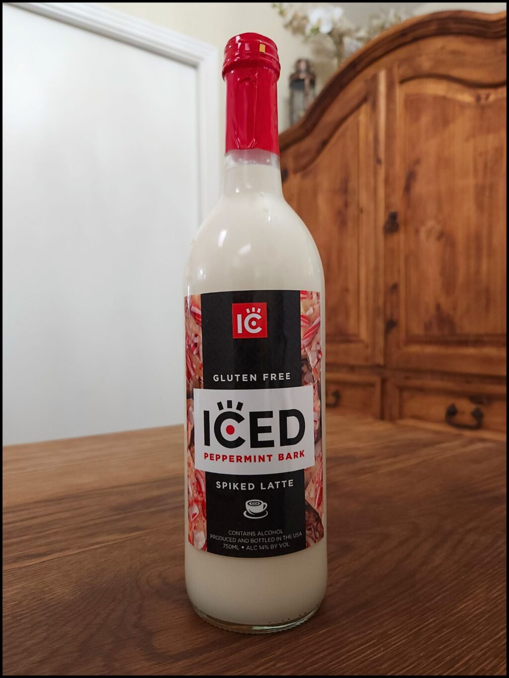 Bottle of IC Iced Peppermint Bark Spiked Latte sitting on a wooden table, in front of a mixed white and wooden background